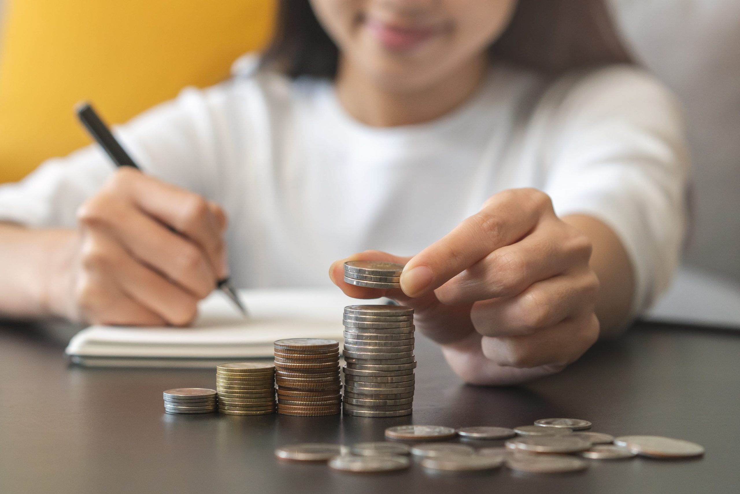 Hand placing a coin on a stack of coins beside a notebook